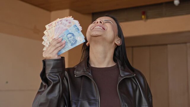 Woman holding and fanning mexican pesos with hand by her face, head tilted back and laughing while wearing a leather jacket inside a building; joy prosperity.