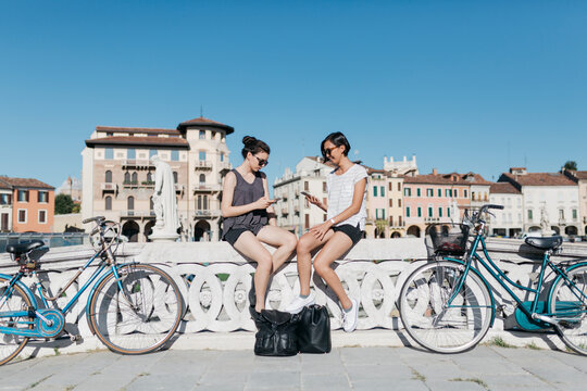 Italy, Padua, two young tourists sitting on railing looking at cell phones