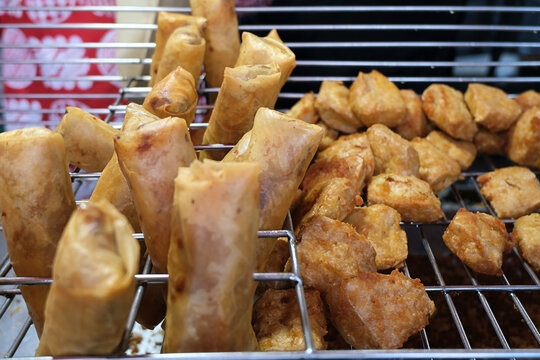 Variety of fried Indonesian snacks,  Lumpia (spring rolls) and Tahu Goreng (fried tofu), arranged on a metal cooling rack highlight a popular Indonesian culinary street food scene for culinary content
