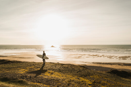 France, Bretagne, Finistere, Crozon peninsula, man standing at the coast at sunset with surfboard