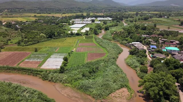 High-angle drone shot tracks over a winding muddy river loop in a lush valley. Cultivated crop fields, patches of agricultural land, and modern greenhouses rest between the water and a rural village.