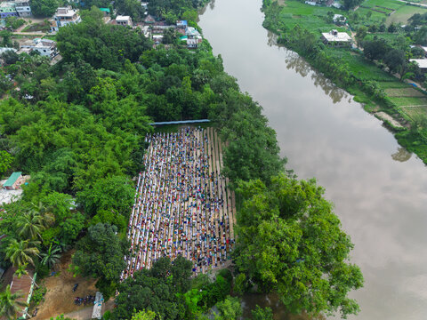 Joypurhat, Bangladesh - 28 May 2026: Aerial view of a large crowd of people gathered for prayer in an open field surrounded by dense green trees and a winding river.