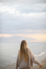 Fototapeta premium Blonde woman standing on seashore at sunset with snowy mountains in the background, warm emotional mood, self care, psychology, nature connection and vacation aesthetic concept