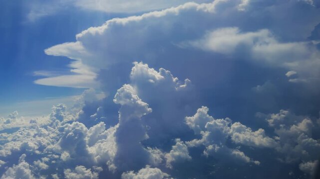 Massive white cumulus clouds billow and tower against deep blue sky as seen from airplane window at high altitude. Sunlight illuminates fluffy cloud formations creating dramatic atmosphere.