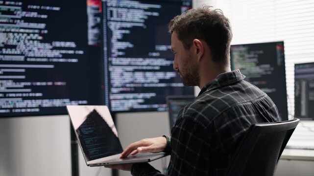 Rear view of computer programmer or hacker using laptop to check server network security system and cyber data privacy code on multiple screens in office workspace.