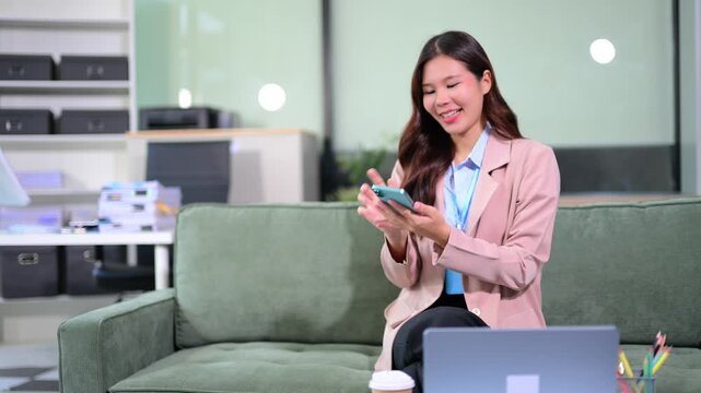 Smiling Asian businesswoman working with laptop, taking notes, and talking on smartphone while sitting on an office sofa.