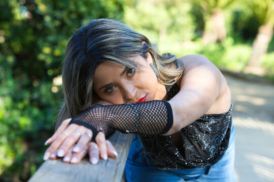 A young, beautiful woman, wearing make-up and dressed in jeans, gloves and a black T-shirt, leans her arms and face against the railing of a wooden bridge and gazes sensually at the camera.