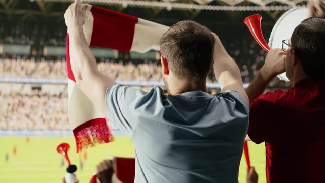 Back view of soccer supporters in red jerseys watching the field from the stands, waving scarves during live football match celebration and team spirit, active supporters chanting on terrace.