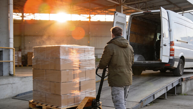 Worker moves boxes with pallet jack to delivery van at warehouse during sunrise