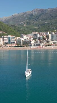 Scenic aerial view of a white sailboat anchored in turquoise adriatic waters off rafailovici, montenegro, with sandy beach, coastal town and mountains under clear skies