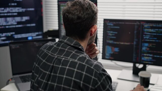 Rear view of thoughtful software developer with hand on chin looking at computer screen, working on server network security system and writing code at an office desk.