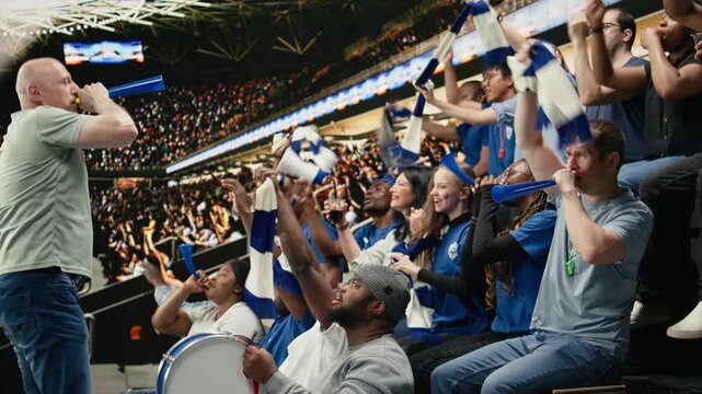 Energetic male football fan blowing blue vuvuzela in stadium terrace while supporters cheer during exciting live soccer match, fan leader and hardcore supporters wearing face paint, stadium noise.
