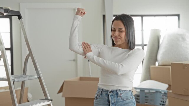 Woman flexes arm and holds elbow while smiling among moving boxes and ladder in building; confidence moving day.