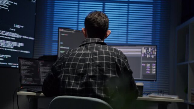 Back view of professional web developer coding software on computer, male application programmer working late at night on internet site development.