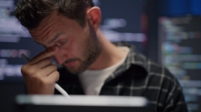 Stressed male web developer rubbing face with headache and fatigue, tired application programmer holding pen stylus and working late on computer code at night.