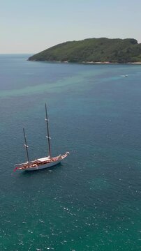 Aerial view of a beautiful white two-masted yacht sailing on the turquoise sea near a green island. Sunny summer day perfect for a vacation