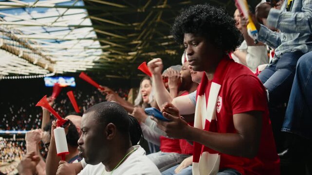 Black sports fan placing football betting wager through mobile sportsbook during live soccer match with cheering supporters at the stadium, man experiencing sports betting tension and anticipation.