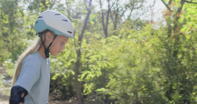 Blonde girl rolls along park path wearing helmet and protective pads. Child keeps arms ready for balance gliding forward on inline rollerblades with steady movement