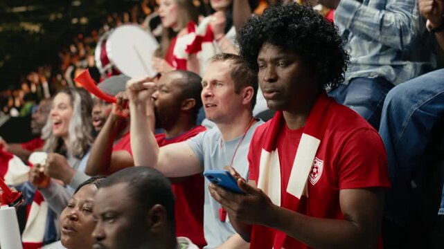 Black sports fan placing football betting wager through mobile sportsbook during live soccer match with cheering supporters at the stadium.