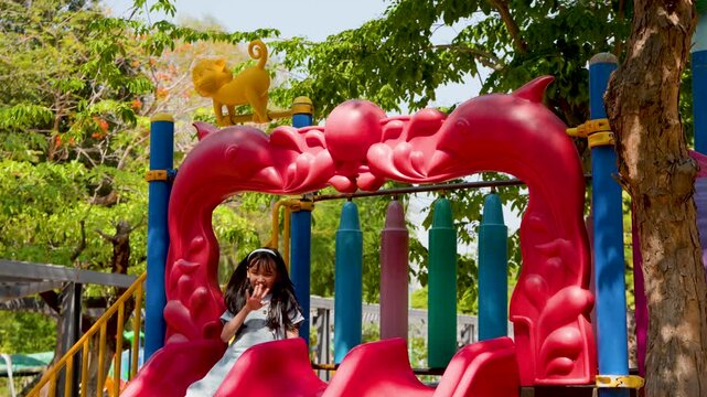 Young Thai Girl in Denim Overalls Waving on Red Playground Slide