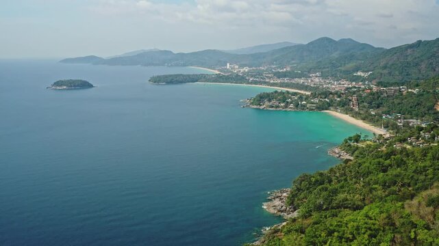 Aerial panoramic flight over Phuket coastline showcasing Karon and Patong beaches, turquoise sea, lush green mountains and resort lined shore creating a serene tropical vacation scene. Travel Thailand