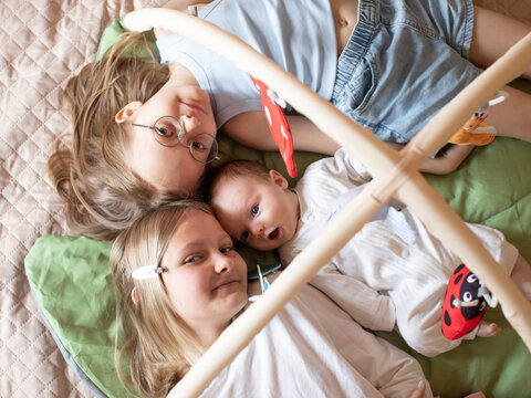 A loving moment with siblings and a baby lying together on a soft surface indoors, older sister with glasses gazing toward the camera while the baby and younger sibling rest nearby, warm natural light