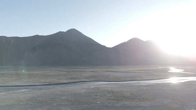 Drone pulls back over a wide barren Ladakh landscape as warm sunlight pushes through gaps between mountain ridges casting a golden glow across the dry valley floor and lighting the dramatic layered.