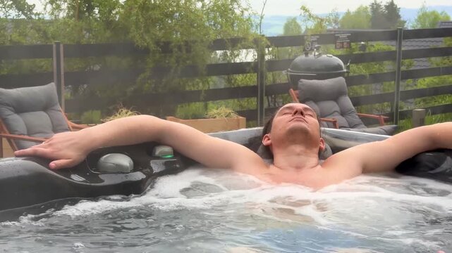 Man relaxing with arms outstretched in hot jacuzzi overlooking mountain valley and lounge area during rainy weekend getaway. Wellness and peaceful spa lifestyle concept outdoors.