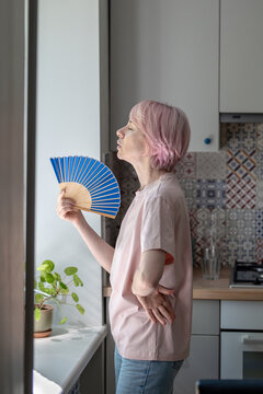 Weary middle aged woman standing in kitchen by window use handheld fan for relief from intense heat. Heatwave, no air conditioning, stifling indoor temperature. Female coping with high warmth at home.