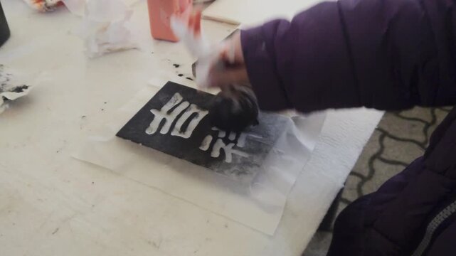 Outdoor static close up shot captures a participant using a round ink dabber tool to apply black ink evenly onto a carved wooden calligraphy printing matrix at a street festival workshop booth.
