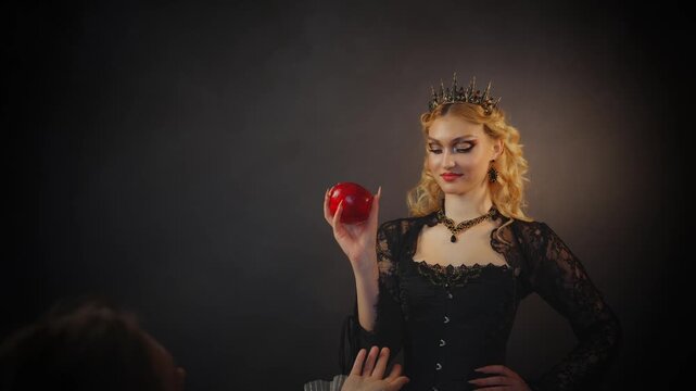 Fantasy fairy woman holding ripe red apple in hands, blonde hair evil queen hand offers poisoned enchanted apple gift to young beauty princess . Royal victorian black dress two women gothic person.