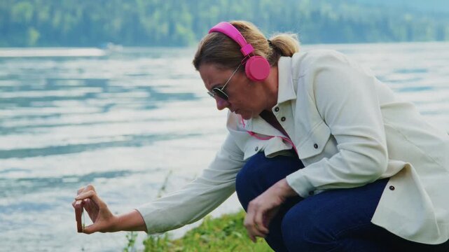 Woman crouches by lake, holding phone to capture nature. Wears pink headphones, sunglasses, focused on water. Relaxed, peaceful, enjoying outdoor moment. Perfect for travel, wellness.