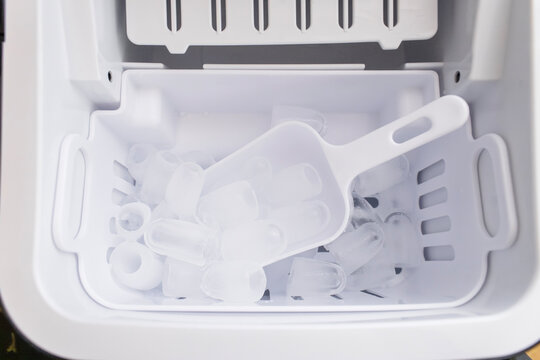 Overhead view inside modern white ice maker showing basket filled with clear bullet ice cubes and plastic scoop. 