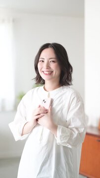 [スローモーション]スマートフォンを胸に抱えて持つ笑顔の女性のライフスタイルとカメラ目線で笑顔のコミュニケーションイメージ縦動画