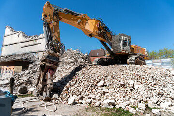 Fototapeta premium A yellow crawler excavator with hydraulic demolition attachment works on a pile of broken concrete, brick rubble, and construction debris beside a partially demolished building under clear blue sky.