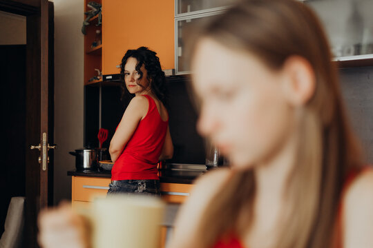 Teen daughter blurred in foreground with distant look while mother watches from kitchen background evoking silent tension in family scene with emotional undercurrent subtle intergenerational conflict