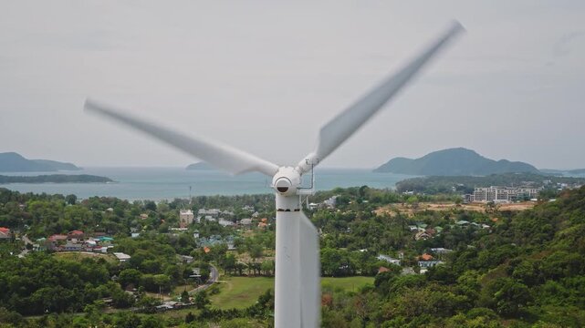 Close up wind turbine blades rotating fast, generating renewable clean energy for the local community, standing prominently against the coastline and ocean on Phuket island. Aerial view drone shot