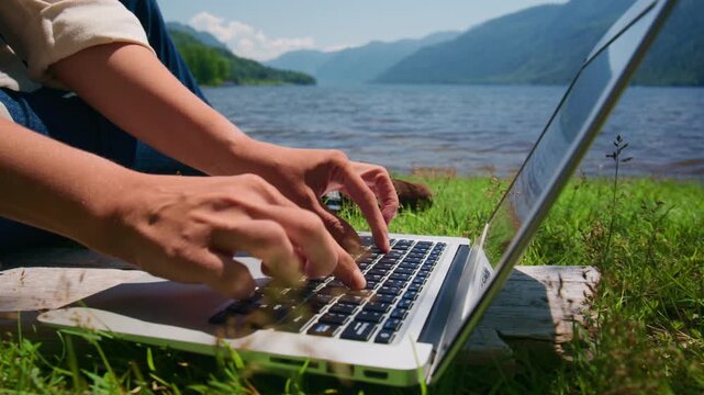 Person types on laptop beside calm lake, green hills behind. Hands move fast, focused on screen. Peaceful outdoor work scene, natural light. Ideal for remote work, travel, or digital nomad themes