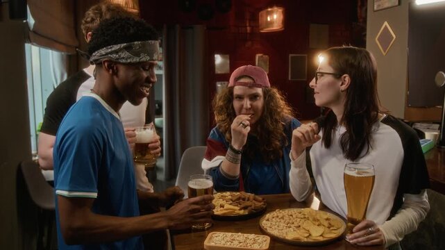 Handheld shot of cheerful young multiethnic friends wearing different team jersey having lively discussion over sports match result while enjoying beer and snacks in bar