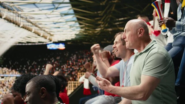 Frustrated football supporter watching tense match moment from stadium stands while crowd behind him cheering with scarves.