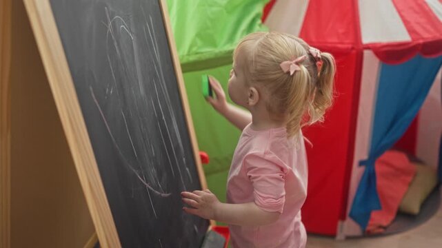 Little blonde girl erases a chalkboard with a green eraser then arranges colorful puzzle pieces on a table in a preschool; curiosity learning.