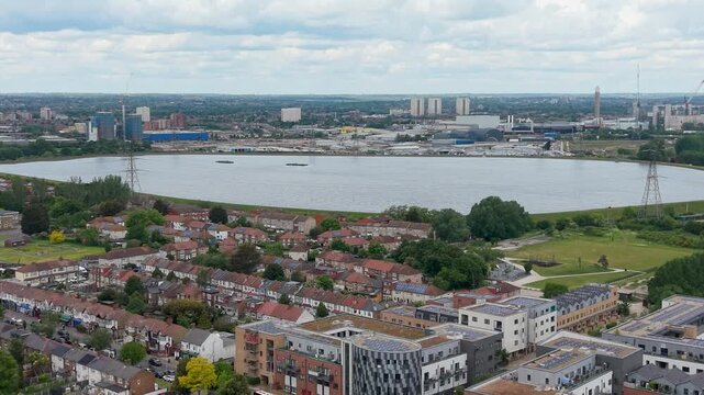 Banbury reservoir drone pullback toward Walthamstow from South Chingford, showing rooftops, parkland, Lea Valley water, pylons, and distant London skyline under clouds.