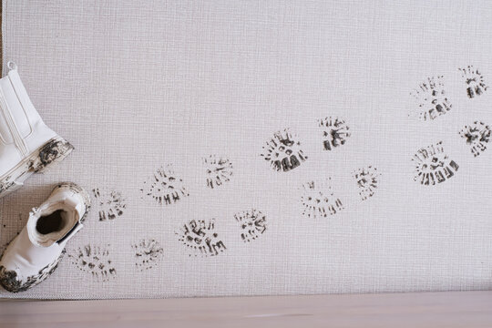 Dark mud-covered footprints lead across a light rug. In the left corner, there are white boots stained with mud after walking outside in rainy or wet weather. top view