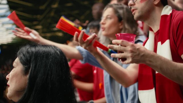 Red team football supporters reacting with anxious gestures in stadium stands during tense live soccer match, scarves and crowd emotion.
