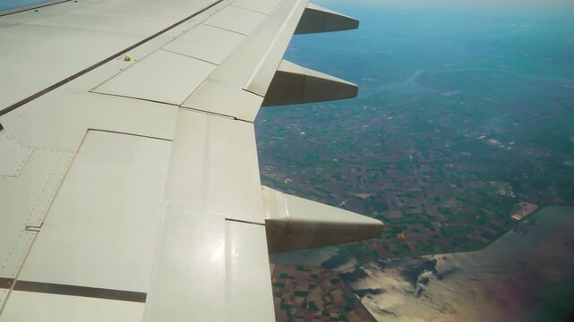 Plane flying above patchwork fields and coastline with visible spoilers. Jet cruising over colorful farmland and river with wing structure in frame. Airliner gliding across mixed landscape showing