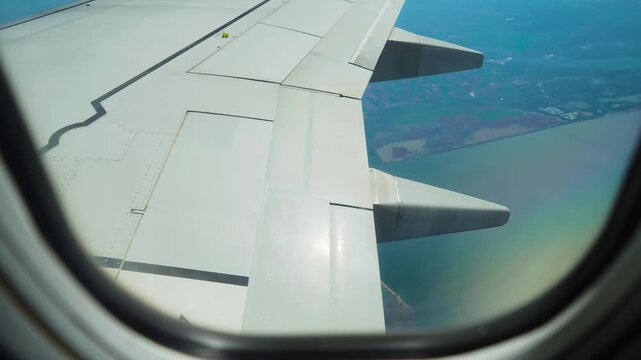 Plane flying above coast with farmlands and industrial zones near water, view from window porthole. Jet cruising over shoreline with mixed terrain and urban edges. Aircraft gliding across transitional