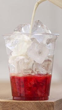 Process of making a strawberry matcha latte with fresh mashed berries. Milk is poured into a plastic cup with strawberry puree and ice cubes.