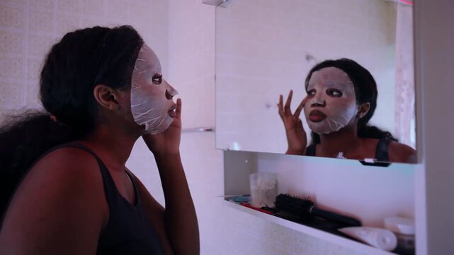 African American woman wearing facial sheet mask looking into bathroom mirror, adjusting skin treatment with focused expression, personal care routine and wellness moment at home