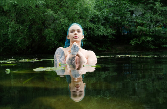 Blue haired tattooed woman standing in dark reflective water surrounded by forest, creating a mysterious cinematic atmosphere of silence and strength