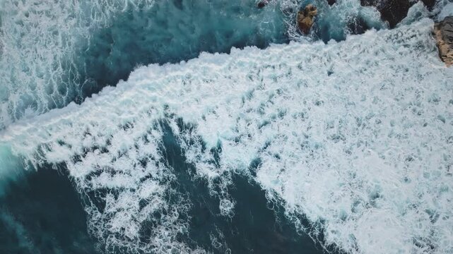 Aerial view discovering powerful turquoise ocean waves breaking with white foam patterns, creating a dynamic and abstract texture along the rugged coastline of Green Bowl Beach. Top down drone shot